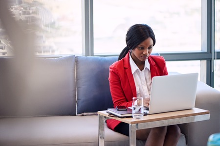 Attractive businesswoman wearing a red blazer busy working on her notebook computer in a business lounge, seated on a blue and grey couch with foreground blur visible in the image.の写真素材