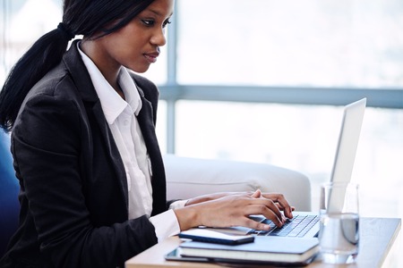 Young adult african business woman wearing a black outfit, busy looking at screen while typing on her notebook, while seted on the sofa in a modern business lounge with large windows.の写真素材