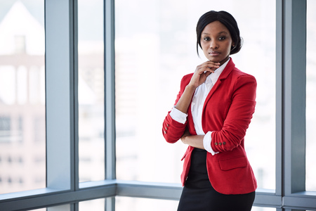 Confident businesswoman looking at the camera with bold body language while wearing a red blazer with large windows behind her in the background.の写真素材