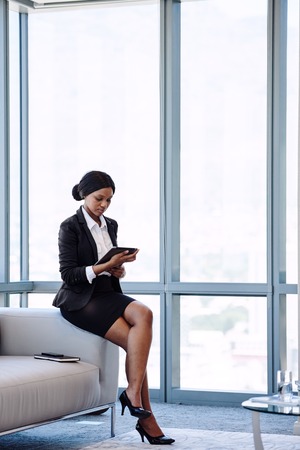 Young black businesswoman sitting on the edge of a sofa in a business lounge while looking at the screen of the digital tablet that she is holding in her hands.の写真素材