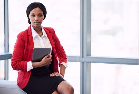 Confident african american businesswoman holding a digital tablet while looking confidently into the camera while seated on the edge of a sofa in a formal business lounge in front of large windows.の写真素材