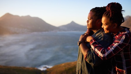 Interracial couple of african descent holding one and other while looking sideways towards the beautiful view of the ocean and mountains in the distance behind them.の写真素材