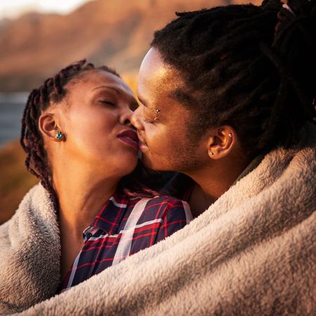 Close up square image of mixed race couple kissing with a blanket wrapped around them to keep warm outdoors, with a view of the ocean and mountains in the distance behind them.の写真素材