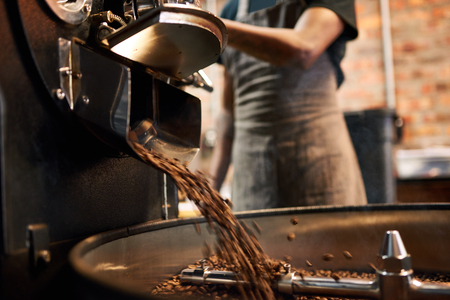 African man wearing an apron busy pouring coffee beans from the coffee roasting machine over to the tray that stirs the beans until they are cool enough to be packaged.の写真素材