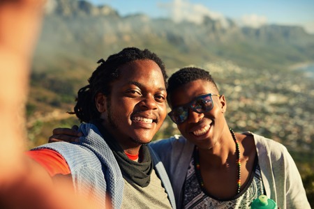 Two happy male african friends take a selfie together while hiking outdoors with gorgeous mountains in the background and true emotion in their smiles.の写真素材