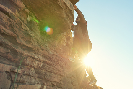 African guy climbing a near vertical rock face with clear blue skies and flare from the sun shining through from behind the climber.の写真素材