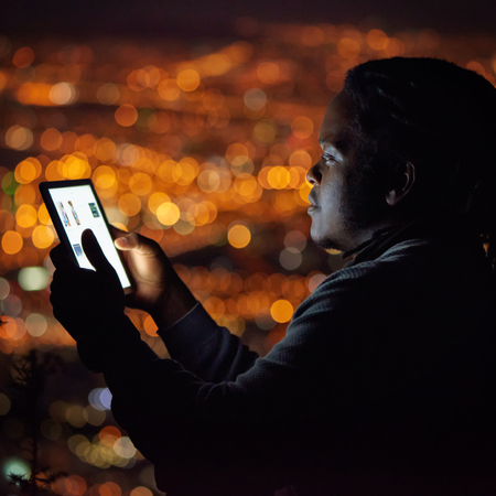 Square image of an African man busy using an electronic tablet with a beautiful bokeh of city lights in the background behind him, and his face illuminated by the light emanating from the screen.の写真素材