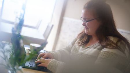 Blurred foreground while chubby white woman sits in a modern cafe with bright light, working on her latest project as she types o her digital tablet.の写真素材