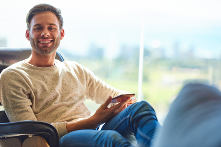 Caucasian man sitting on a modern couch next to large glass windows with a stunning view behind him as he smiles at camera with his phone still being held in his hands.の写真素材