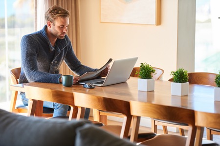Sophisticated modern man sitting at his dining table with his morning cup of coffee at his side and his notebook ready for work in front of him while reading the daily newspaper.の写真素材