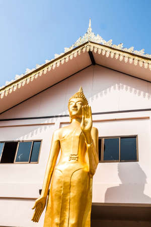 Standing bronze buddha image statue in front of monk school buildingの写真素材