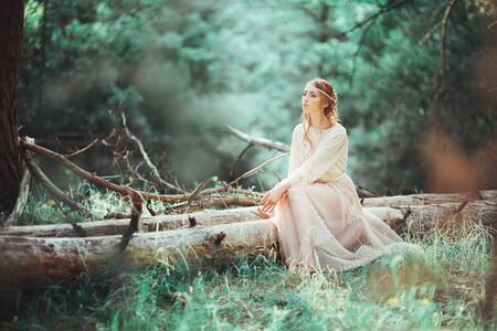 Beautiful artistic photo ginger girl in white dress sitting on a tree in the woods.の写真素材