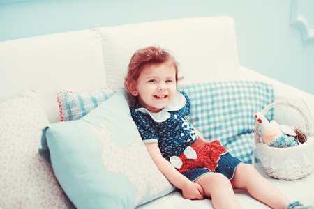 A beautiful little red-haired girl in a dress sitting on a white sofa, laughs and playsの写真素材