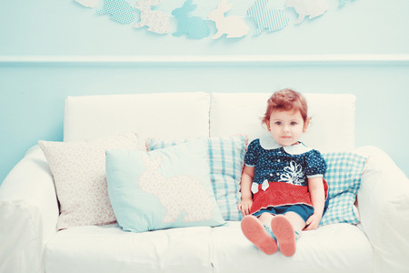 A beautiful little red-haired girl in a dress sitting on a white sofa, laughs and playsの写真素材
