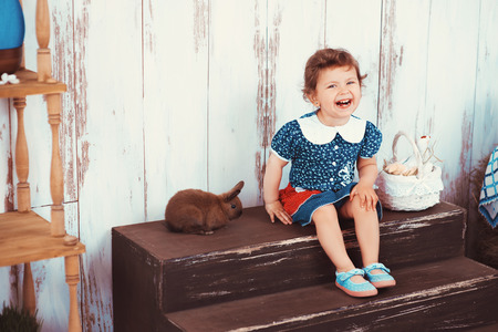 A beautiful little red-haired girl in blue dress with basket of Easter plays in the interior with a rabbitの写真素材