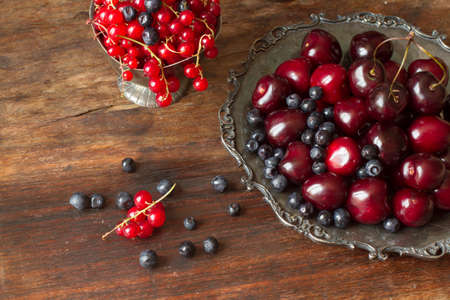 cherry with red currants and blueberries in a jug and a metal plate in oriental style on a background of woodの写真素材