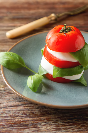 Fresh italian Caprese salad tomato and mozzarella slices with basil leaves on a blue plate with vintage yellow fork. wooden backgroundの写真素材