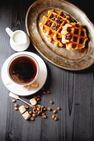 Homemade belgian waffles, white ceramic cup of coffee, milk, teaspoon and coffee beans. Dark rustic background. Space for text in the centerの写真素材