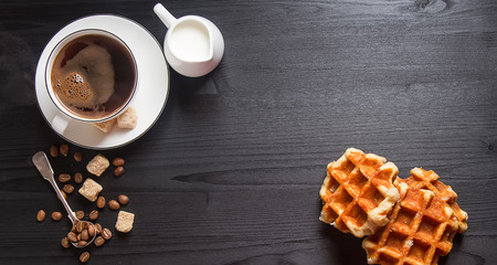 Homemade belgian waffles, white ceramic cup of coffee, milk, teaspoon and coffee beans. Dark rustic background. Space for text in the centerの写真素材