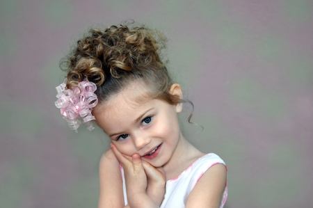 Little girl leans her head in her hands   She has curls all over her head and a sweet curly ribbon bow decorating it   Background is mottled green and pink の写真素材