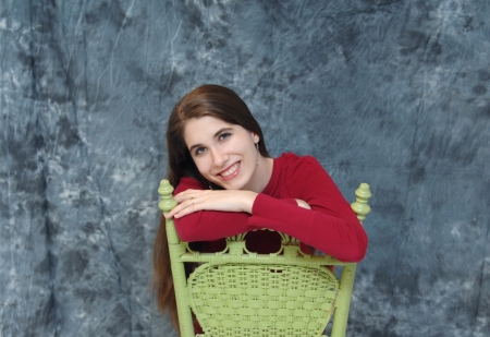 Female teen sits on a green, wooden chair backwards.  She rests her arms on the chair and smiles.  Image has grey blurred background.の写真素材