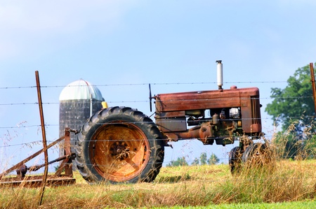 A relic of farming is parked at the fence row to be sold.  Tractor and bushhog stands idle and rusting.  A lone silo stands in the background.のeditorial素材