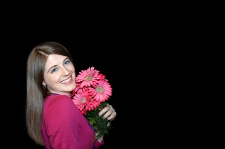 Beaming young woman holds bouquet of pink daisies.  She is clutching bouquet close to her face.  Background is black and subject is in corner for copy space.の写真素材