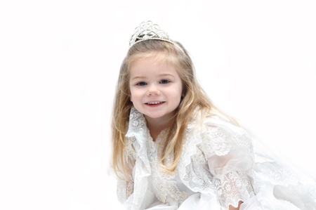 Little girl wears an elegant wedding dress and crown.  She is sitting in an all white room and smiling happily as she dreams of her getting married when she grows up.  の写真素材
