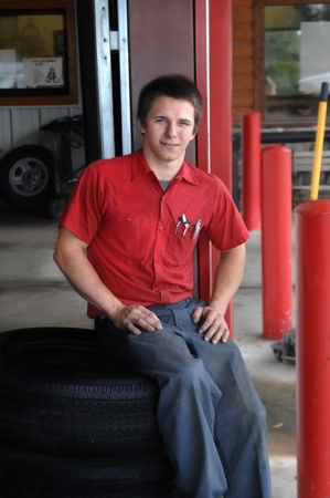 Young man rests on tire at the tire store that he works at   His uniform is red and grey and he is smiling の写真素材