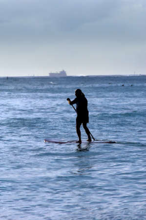 Older man paddle boards across Waikiki Bay while ship is leaving port   Evening light silhouettes paddler の写真素材