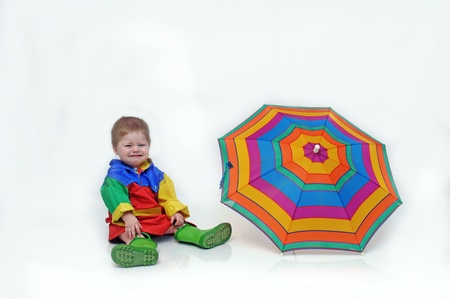 Baby boy, wearing a colorful slicker suit and rubber boots, is sitting besides an open umbrella and crying の写真素材