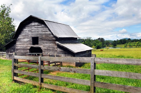 Horse grazes in the pasture close to a distant pond   Barn and rustic wooden fence are both weathered and worn   Blue sky and clouds make for perfect country scene の写真素材