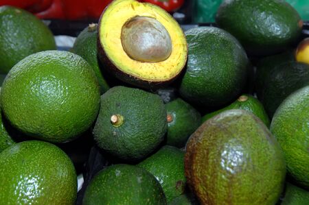 One avocado, in a pile of twenty or more, has been cut in half to show the fruit pulp and seed 