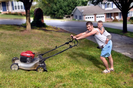 Little boy rides piggy back while dad mows the family's front yard.  Dad is grimacing and child is smiling happily.の写真素材