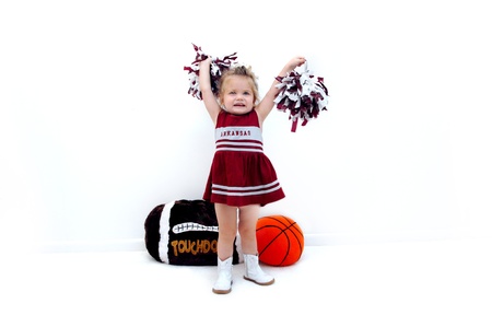 Little cheerleader dressed in burgundy and grey holds pom poms high in the air   She is wearing white gogo boots and standing in front of a stuffed football and basketball の写真素材