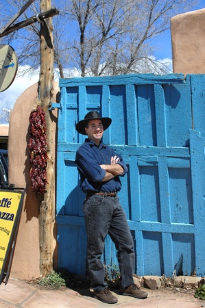 Man wearing a black leather hat and a big smile, poses besides a vivid blue gate in the town of Taos, New Mexico   Gate is wooden, rustic and leaning   