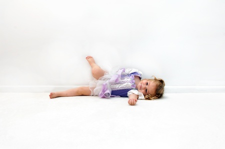 A tired little ballerina lays on the floor in an all white room.  She is dressed in a ballerina costume of lilac and purple.  Child is grinning and one foot rests on the wall.の写真素材