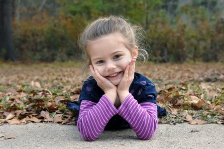 Adorable little girl lays on the Fall leaves and leans her head in her hands   She is smiling and...