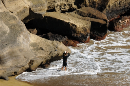 Young male dons his hat in preparation for fishing the famous Green Sand Beach on the Big Island of Hawaii.  Huge cliff is rugged and eroded by wind and waves.  Size of cliff is seen in comparison to the small stature of man.の写真素材