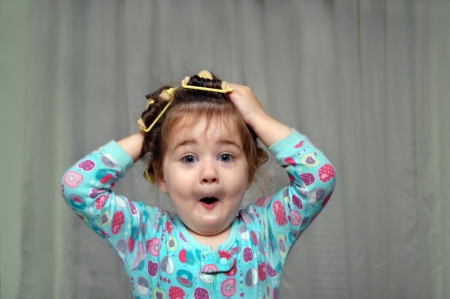 Old fashioned sponge curlers make this little girl want to pull them out.  She is making a comical face as she puts her hands on her head.の写真素材