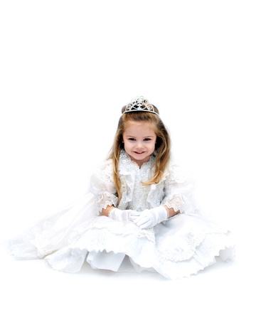 Wishful little girl is dressed up as a bride with sparkling crown tiara   She is smiling happily and sitting in an all white room の写真素材
