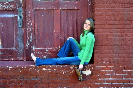 Attractive young woman sits in front of alley doorway   Paint is peeling and cracked   She is wearing jeans and is barefoot の写真素材