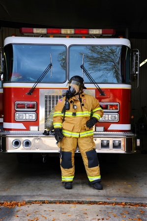 Woman firefighter stands in front of fire truck at fire station.  She is wearing bunking gear complete with gas mask and helmet.の写真素材