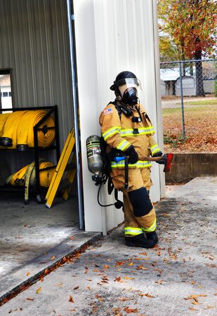 Fire Woman stands holding flat head axe as she leans against fire station.  She is suited up in her Bunking Gear and is ready for any fire emergency.の写真素材