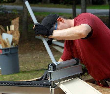 Young construction worker uses equipment to cut pieces of siding for home improvement project.  He is wearing a red T-shirt and wearing a tool belt, hat and safety glasses.の写真素材