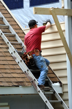 Young carpenter remodels aging home.  He is installing siding and is using a level to be sure installation is precise.  He is standing on a ladder on rooftop.の写真素材