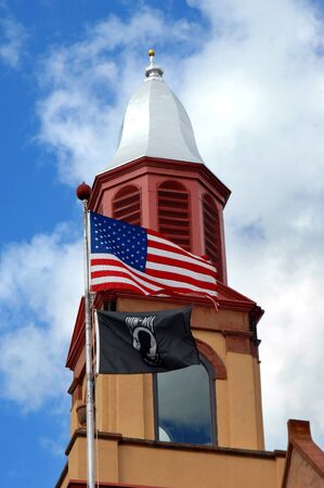 Octagonal cupola and finial, on the Lake Linden Village Hall and Fire Station, is framed by blue sky and clouds.  An American flag and POW flag fly on pole besides it.のeditorial素材
