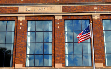 American flag flies in front of a high school building.  Windows reflect clouds and blue sky and the words "high school" is engraved on the front of building.のeditorial素材