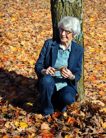 Elderly woman leans against a tree and is surrounded by the leaves of Autumn in North Arkansas.  She is wearing jeans and jean jacket.の写真素材