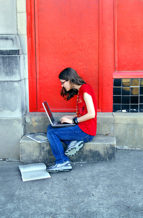 Young high school student holds a lap top as she sits on the steps of her school.  She has her books at her feet and is doing research on a homework paper.の写真素材